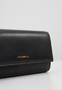 Portefeuille en cuir noir texturé avec le logo doré "Coccinelle" centré sur le rabat, posé sur une surface claire unie.