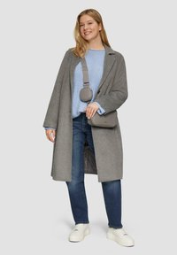 Grauer Wollmantel über einem hellblauen Strickpullover, blaue Jeans und weiße Sneakers. Eine graue Umhängetasche hängt an einem Riemen.