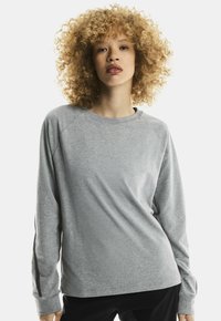 Young person with curly blond hair wearing a light gray long-sleeve shirt and black pants against a plain background.