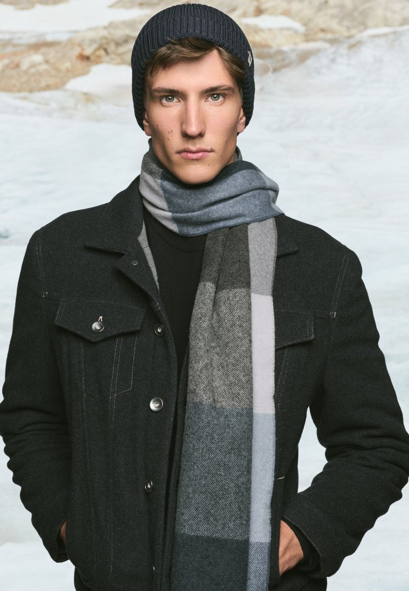 Black wool blend coat with silver buttons, black turtleneck, and grey plaid scarf, featuring shades of grey and blue. Dark knit beanie on head.