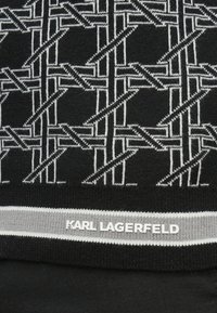 Juodos megztos medžiagos su baltu geometriniu raštu. Pilka juosta apačioje turi baltomis raidėmis užrašytą „KARL LAGERFELD“ etikėtę.