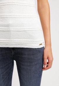 Top bianco con texture e motivi a righe orizzontali, accompagnato da un piccolo etichetta con marchio, abbinato a jeans di denim scuri. Cuciture dettagliate visibili.