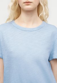 Νεαρός με ανοιχτόχρωμα σγουρά μαλλιά που φοράει ένα απλό ανοιχτό μπλε κοντομάνικο μπλουζάκι μπροστά σε απλό φόντο.