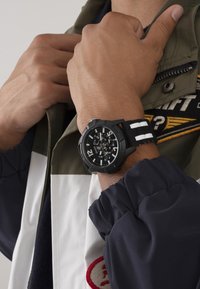 Černé chronografické hodinky s bílými pruhy na gumovém pásku, obsahující velký kulatý ciferník a detailní displeje, nošeny na zápěstí.