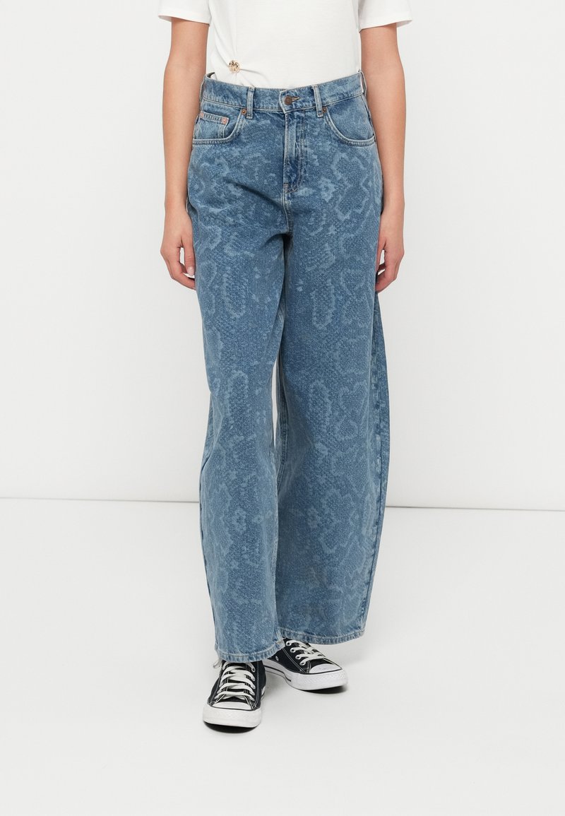 Lichtblauwe wijde jeans van denim met een subtiel slangenprintpatroon, vijf zakken en een klassieke knoopsluiting, gecombineerd met zwarte sneakers.