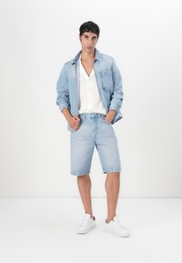 Lys blå denimjakke og shorts, paret med en hvit strikket skjorte og hvite joggesko. Antrekket har en avslappet passform og klassisk snitt.