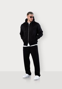 Schwarze Zip-Hoodie mit geprägtem Schriftzug, kombiniert mit schwarzen Hosen. Darunter ein weißes T-Shirt und weiße Sneakers, getragen von einer Person.