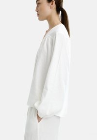 Biała lniana koszula o luźnym kroju, szerokimi rękawami i dekoltem w kształcie litery V. Posiada delikatną teksturę oraz kieszenie po bokach.
