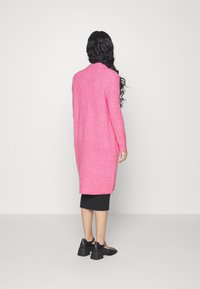 Kvinna med långt mörkt hår som bär en ljusrosa lång stickad kofta, svart midikjol och svarta klackskor med blockklack, som står mot en vit vägg.