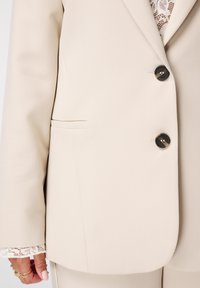 Close-up van een beige blazer met twee donkere knopen, kant manchet details en een hand die gouden ringen en lichtroze nagellak draagt.