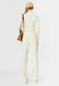 Chaqueta crema clara con cremallera y pantalones acampanados a juego, hechos de tela suave con un brillo sutil. Los accesorios incluyen una gorra beige y un bolso marrón.