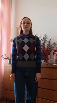 Donna con lunghi capelli rossi che indossa un maglione fantasia e jeans, in piedi all'interno vicino a una cassettiera di legno con composizioni floreali rosse e rosa.