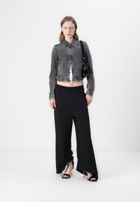 Giacca di jeans grigia tagliata con colletto, zip anteriore e due tasche laterali, indossata su un top bianco corto. Pantaloni neri a palazzo fluenti e tacchi neri.