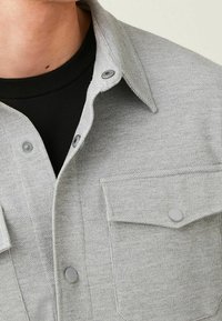 Camicia grigia con bottoni, realizzata in un tessuto testurizzato. Presenta un colletto, due taschini sul petto e bottoni a pressione in una tonalità di grigio abbinata.