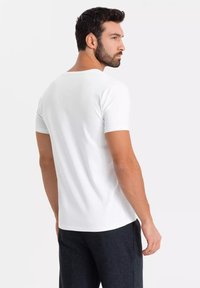 Mężczyzna z ciemnymi włosami i brodą, ubrany w prostą białą koszulkę z krótkim rękawem i ciemne spodnie, stoi i patrzy w bok na tle jednolitego tła.