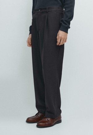 Pantaloni grigio scuro, ampi e a vita alta, con pieghe frontali e tasche laterali. Abbinati a scarpe di pelle marrone.