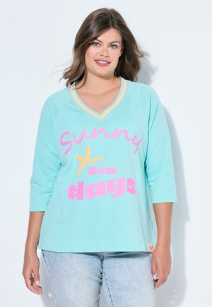 Kvinde med skulderlangt brunt hår har en turkis V-hals skjorte med pink og gul tekst "solrige sødage" og lyseblå jeans på.