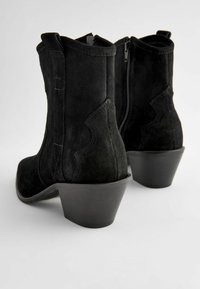 Black suede ankle boots with a curved design, featuring a side zipper and a low, stacked heel. Smooth texture with decorative stitching.