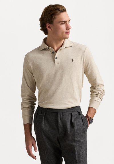 Polo beige à manches longues avec une patte de boutonnage à trois boutons et un logo brodé, associé à un pantalon gris plissé. Texture lisse, design décontracté.