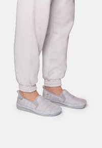 Sive slip-on cipele imaju kviltani dizajn s dlakavim, sivo krznom i teksturiranim sivim potplatom, u kombinaciji s svijetlosivim jogger hlačama.