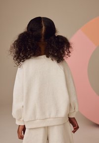 Sweat-shirt blanc texturé avec manches bouffantes et poignets côtelés, porté par un enfant, assorti à un pantalon. Fond pastel doux.