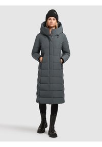 Grijze lange pufferjas met capuchon, voorzien van een doorgestikt ontwerp, zijzakken en een rechte silhouet. Gecombineerd met zwarte leggings en laarzen.