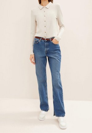 Kobieta ubrana w kremowy, prążkowany kardigan na guziki, niebieskie dżinsy z wysokim stanem i brązowym paskiem oraz białe trampki, stojąca z jedną ręką w kieszeni.