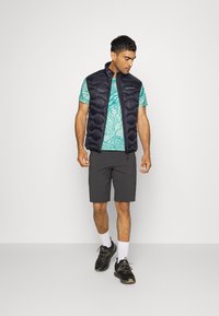 Jūras flotes pūkaini veste pāri tirkīza rakstainai t-kreklam, kopā ar tumšiem šortiem un melniem sporta apaviem. Modelis stāv uz vienkārša fona.