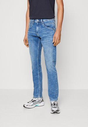 Vaqueros de mezclilla azul claro con un corte recto, que presentan un diseño de cinco bolsillos, cierre de cremallera y un ligero desgastado. Se llevan con zapatos negros y una camiseta oscura.