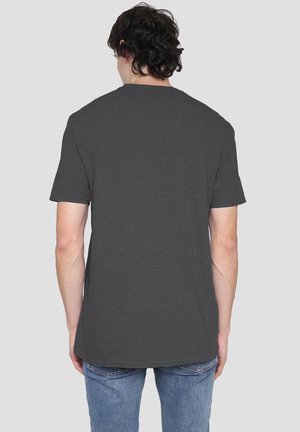 Mann mit dunklen, lockigen Haaren, der ein schlichtes, dunkelgraues Kurzarm-T-Shirt und blaue Jeans trägt und mit dem Rücken zu einer hellgrauen Hintergrund steht.