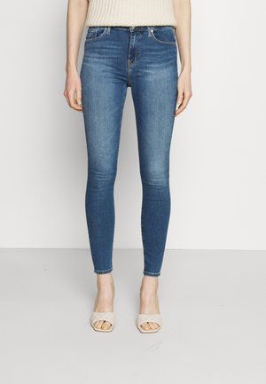 Femme portant un jean skinny bleu et des sandales beiges à talons ouverts, debout sur un fond blanc uni.