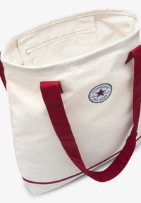 Τσάντα ώμου από καμβά σε κρεμ χρώμα με αντίθετες κόκκινες λαβές, διαθέτει εσωτερική τσέπη με φερμουάρ και λογότυπο Converse All Star στο πλάι.