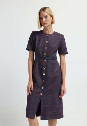 Femme aux cheveux blonds courts portant une robe violette au genou à manches courtes avec des boutons dorés et une ceinture bleu marine, se tenant devant un fond uni.
