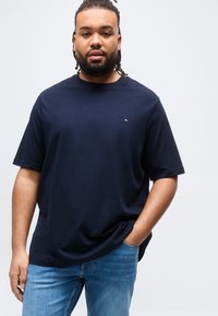 Άνδρας με γενειάδα και κοτσιδάκια φοράει μπλουζάκι σκούρου μπλε και μπλε τζιν, πόζαροντας με το ένα χέρι στην τσέπη μπροστά σε απλό γκρι φόντο.