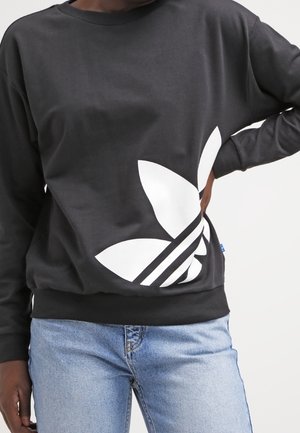 Person iført en sort sweatshirt med et stort hvidt Adidas trefoil-logo og lyseblå jeans, hånd hvilende på hoften.