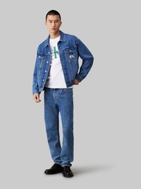 Denim jacket in medium wash with two front pockets and buttons, worn over a white T-shirt. Light blue straight-leg jeans and black shoes.