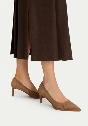 Piernas con zapatos de tacón medio de punta puntiaguda de ante marrón combinados con una falda midi marrón con una abertura frontal.