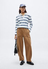 Lange mouw gestreept shirt in wit en marineblauw, bruine broek met brede pijpen, zwarte schoenen en een marineblauwe pet. Zwarte tas met riem. Getextureerde materialen.