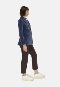 Blazer doppiopetto blu navy in tessuto morbido con bottoni dorati, abbinato a pantaloni corti marrone scuro e sneaker bianche con plateau.