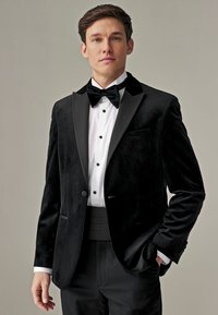 Joven con esmoquin de terciopelo negro, pajarita, camisa blanca de vestir y faja, de pie contra un fondo neutro y liso.