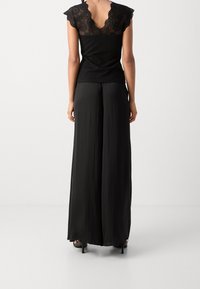 Černý top s krajkovými rukávy ve tvaru čepice, s vroubkovaným vzorem. Kombinován s volnými černými kalhotami. Hladká struktura s elegantními detaily.