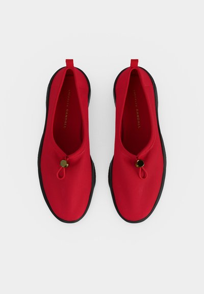 Κόκκινα παπούτσια slip-on με μαύρες σόλες και στρογγυλά χρυσά κουμπώματα από πάνω, σε λευκό φόντο, φωτογραφημένα από πάνω.