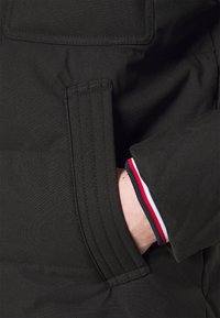 Dunkelgrüne Jacke mit einer Seitentasche, die horizontale Steppdetails aufweist; der Ärmelsaum zeigt einen Dreifarbenstreifen (rot, weiß, blau).