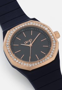 Orologio ottagonale con cinturino in silicone blu navy, lunetta in oro rosa, quadrante nero con texture e dettagli in cristallo attorno al quadrante, con lancette in oro rosa.