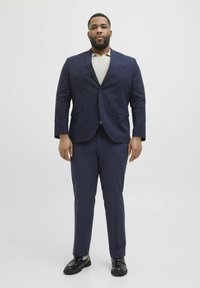 Costume marine en tissu texturé, comprenant une veste à revers et un pantalon assorti, associé à un polo beige clair et des chaussures noires.