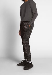 Cargo pants in patterned dark grey with a slight sheen, featuring side pockets. Paired with a fitted dark grey short-sleeve shirt.