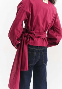 Bordowa bluzka kopertowa z bufkami na długich rękawach, zawiązywana w talii. W zestawie ciemne dżinsy z klasycznymi kieszeniami i szczegółami przeszyć.