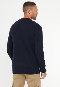 Námořnický modrý pletený svetr s dlouhými rukávy a žebrovanou strukturou, má kulatý výstřih a volný střih, v kombinaci s béžovými kapsáčovými kalhotami.