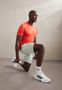 Sportists oranžā īsajā piedurkņu krekls un gaiši zaļās šortos, ceļos noliecies, katrā rokā turot sudraba hanteles, ģērbies baltās sporta kurpēs.