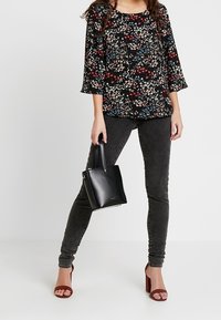 Kvinna som bär en blommig blus, mörka jeans och röda klacksandaler, håller en liten svart handväska och står mot en neutral bakgrund.